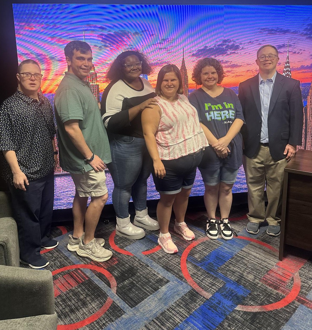 Six people standing together in front of a colorful television studio backdrop